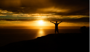 A man reaching his arms out towards the heavens with the sun on the horizon
